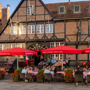 Außenansicht des BLOCK HOUSE Hamburg Bergedorf mit gut besuchter Terrasse, roten Sonnenschirmen und historischem Fachwerkgebäude.