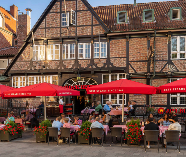 Außenansicht des BLOCK HOUSE Hamburg Bergedorf mit gut besuchter Terrasse, roten Sonnenschirmen und historischem Fachwerkgebäude.