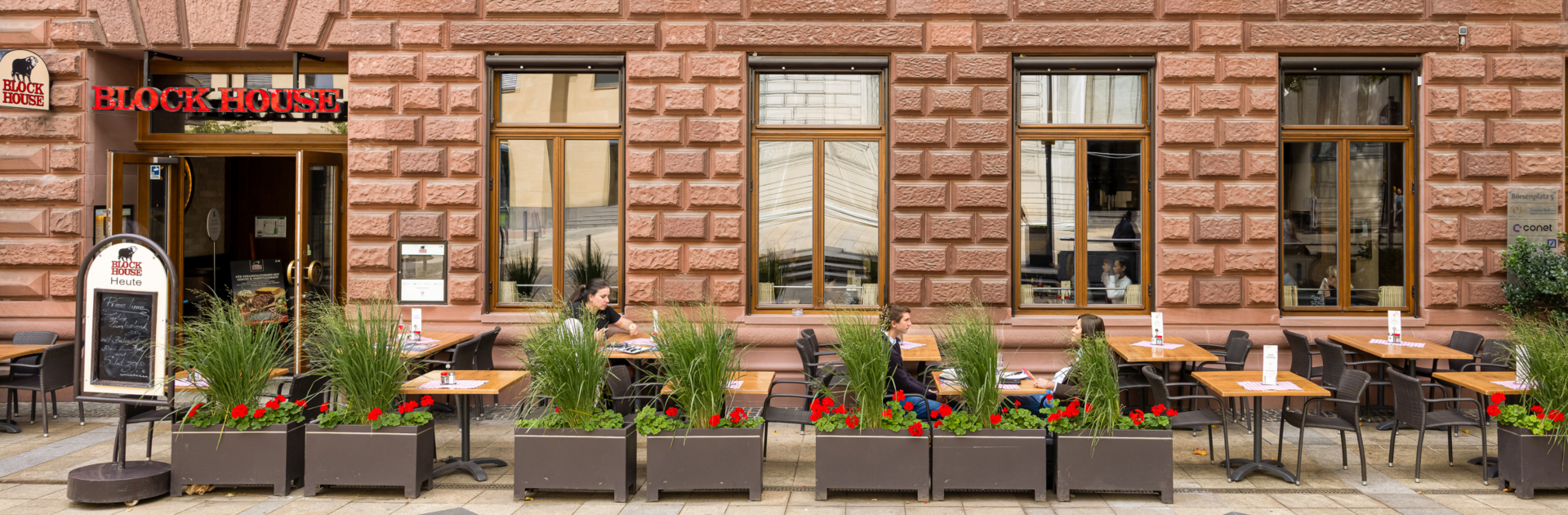 Außenansicht des BLOCK HOUSE Frankfurt am Börsenplatz mit Terrasse, begrüntem Außenbereich und Gästen vor dem Restaurant.