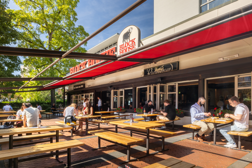 Terrasse des BLOCK HOUSE Frankfurt Westend mit Gästen an Holztischen vor dem Steakhouse unter roten Markisen
