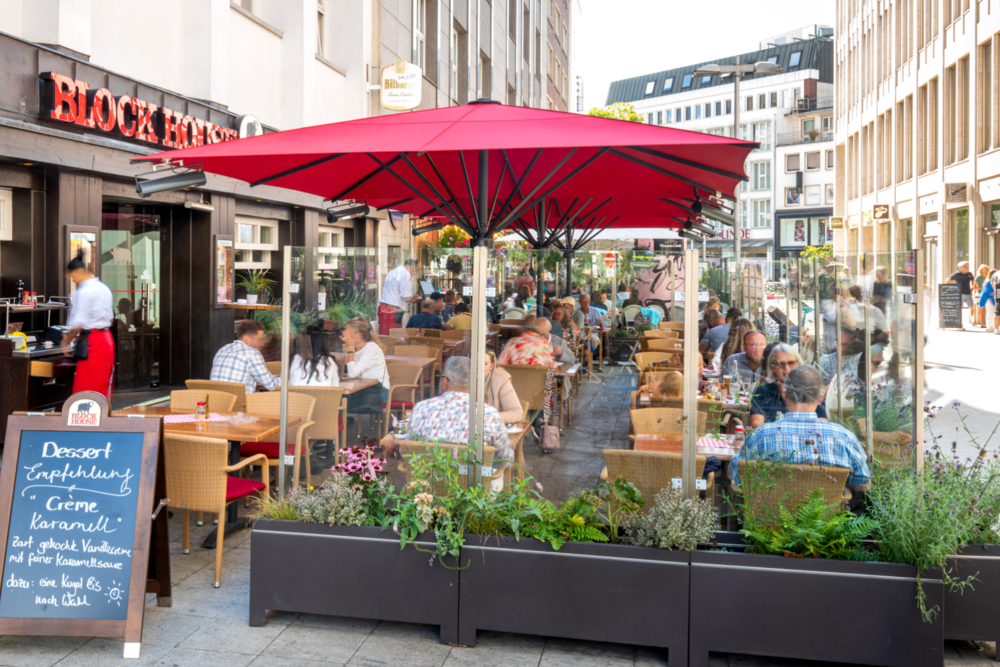 Terrasse des BLOCK HOUSE Hannover Am Kröpcke mit Gästen unter roten Sonnenschirmen in der Fußgängerpassage der Innenstadt