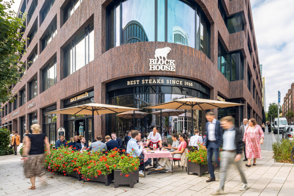 Außenansicht des BLOCK HOUSE Deutschlandhaus mit Terrasse und Gästen vor dem Restaurant.