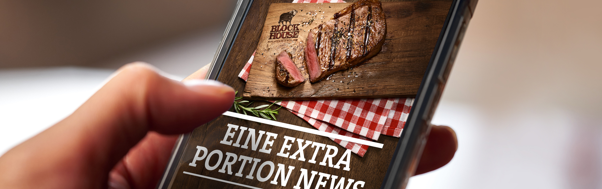Person hält Handy in der Hand, wo ein Teaser des BLOCK HOUSE Website Newsletters mit der Aufschrift "Eine extra Portion News" mit einem Steak zu sehen ist