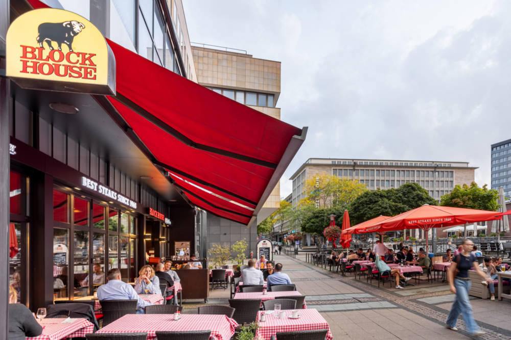 Außenbereich des BLOCK HOUSE Essen mit Terrasse, roten Markisen und Gästen auf dem Platz.