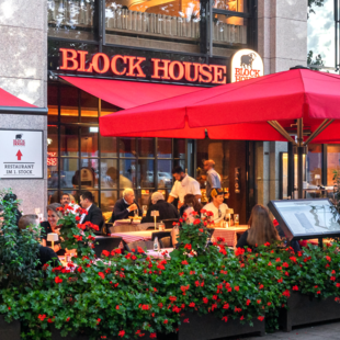 Außenansicht des BLOCK HOUSE München Elisenhof mit Terrasse, roten Sonnenschirmen und Gästen im Außenbereich.