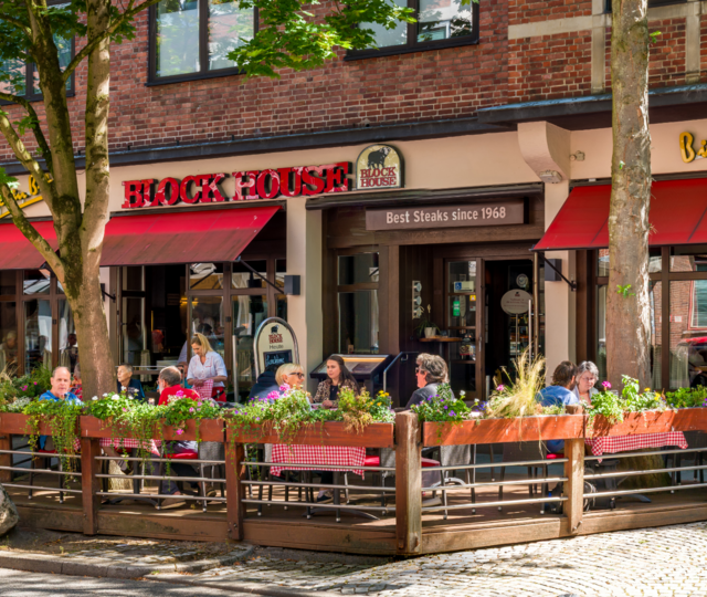 Außenansicht des BLOCK HOUSE Kiel mit Terrasse, roten Markisen und Gästen vor dem Restaurant.