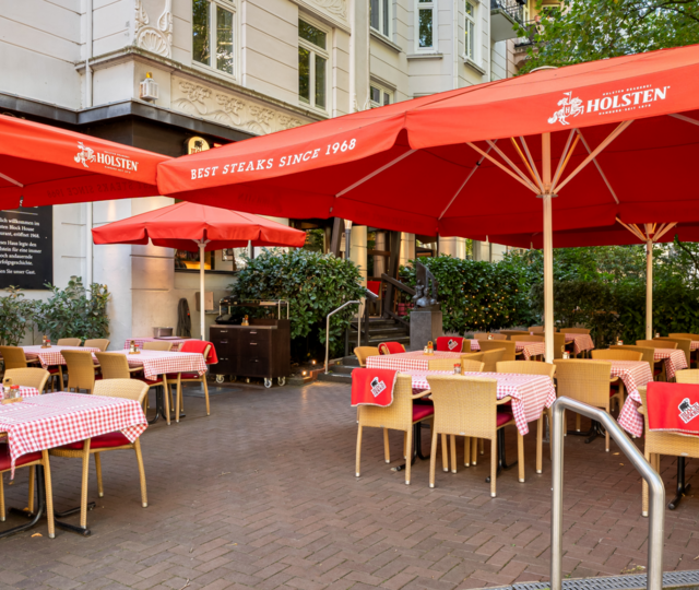 Eingangsbereich und Terrasse des BLOCK HOUSE Hamburg Winterhude mit Sonnenschirmen, Sitzplätzen und altem Baumbestand