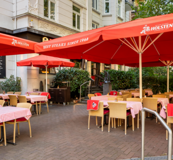 Eingangsbereich und Terrasse des BLOCK HOUSE Hamburg Winterhude mit Sonnenschirmen, Sitzplätzen und altem Baumbestand