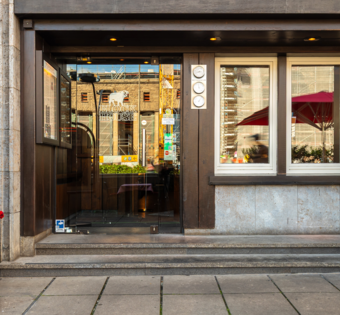 Eingang des BLOCK HOUSE Stuttgart Arnulf-Klett-Platz mit Menütafel vor dem Steakhouse und Gästen an Tischen mit rot-weiß karierten Tischdecken