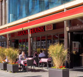 Außenansicht des BLOCK HOUSE Frankfurt Europa-Allee mit ausgefahrener Markise und Gästen auf der Terrasse bei gutem Wetter