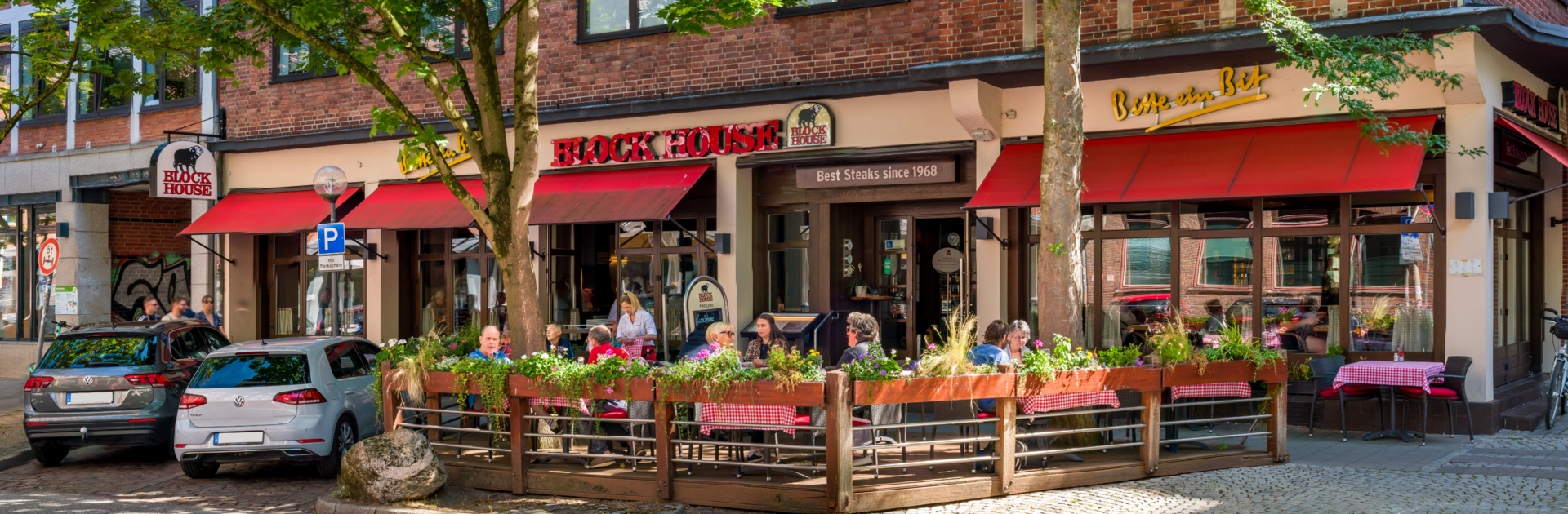 Außenansicht des BLOCK HOUSE Kiel mit Terrasse, roten Markisen und Gästen vor dem Restaurant.