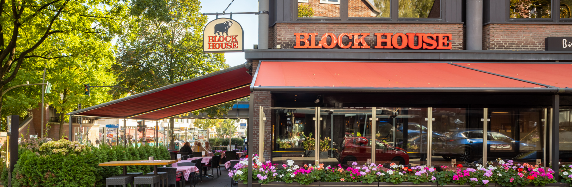 Außenansicht des BLOCK HOUSE Hamburg Barmbek mit Terrasse, roten Markisen und Sitzplätzen vor dem Steakhouse